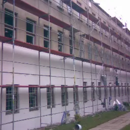 Prace na wysokościach: budynek wielopiętrowy w trakcie docieplania, widoczne rusztowania, pracownicy w białych koszulkach przy pracy, biała elewacja, fragment chodnika i trawnika.